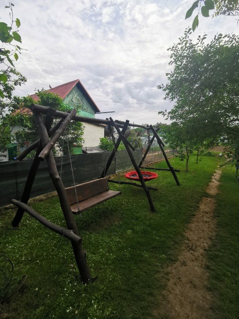 Drewniana huśtawka z siedziskiem na łańcuchach i okrągła huśtawka typu bocianie gniazdo, ustawione w ogrodzie z widokiem na dom z czerwoną dachówką.