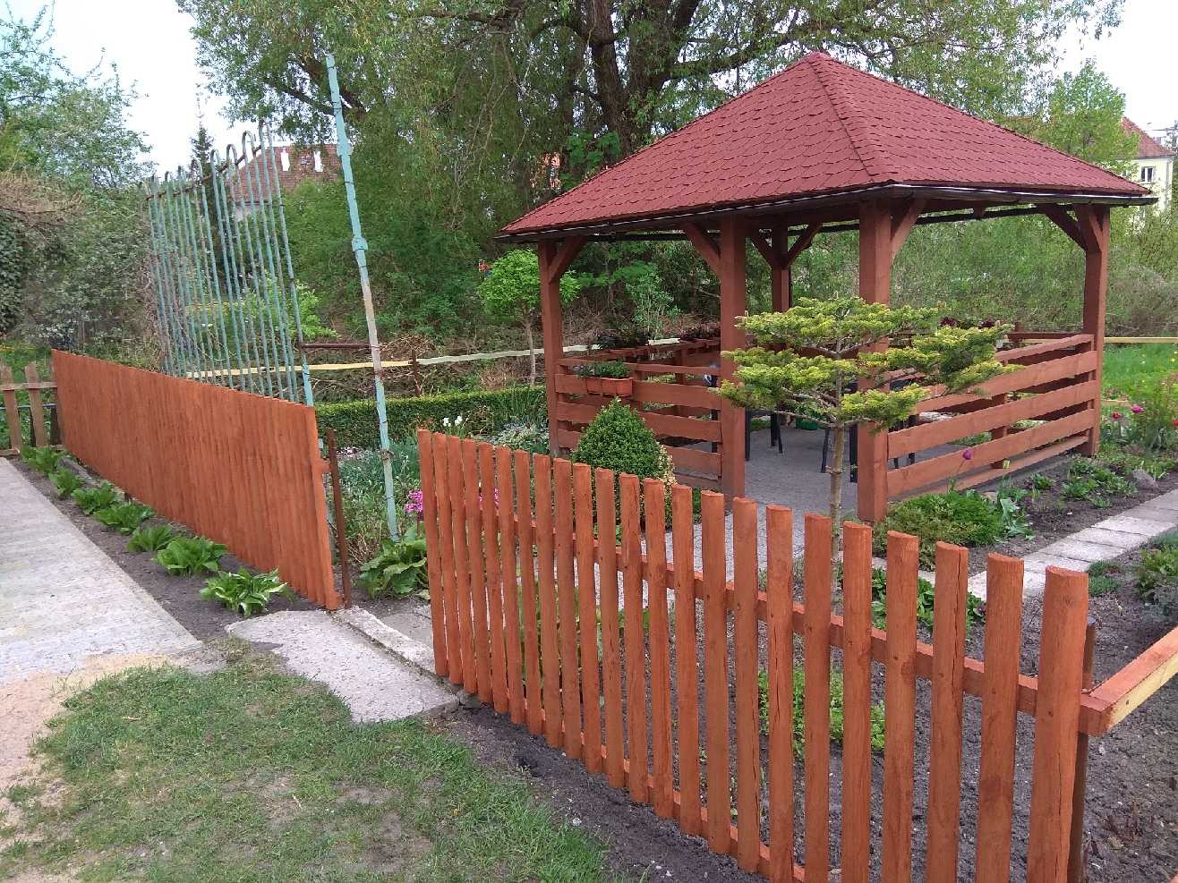 Nowe, drewniane ogrodzenie w kolorze miodowym otacza zadbany ogród z altaną z czerwonym dachem, widok z poziomu trawnika.