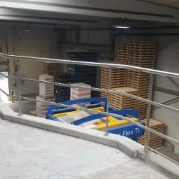 Metalowa balustrada ochronna zamontowana w hali magazynowej, widoczne palety i wózek widłowy.