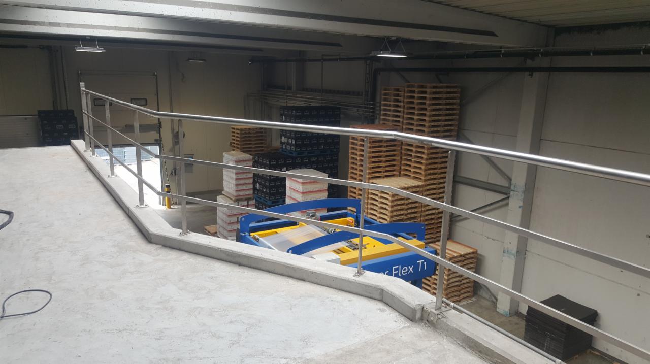 Metalowa balustrada ochronna zamontowana w hali magazynowej, widoczne palety i wózek widłowy.