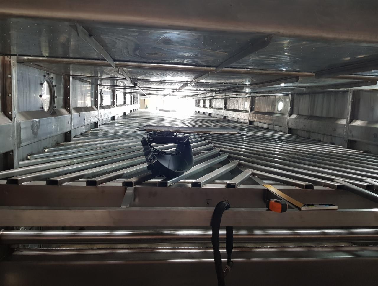 Wnętrze metalowej konstrukcji z widocznymi spawami, maską ochronną, miarką i poziomnicą, oświetlone jasnym światłem w oddali.