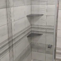 Wnętrze kabiny prysznicowej z trzema półkami narożnymi z szarego kamienia i odpływem liniowym w podłodze wyłożonej płytkami imitującymi drewno, wentylator na ścianie.