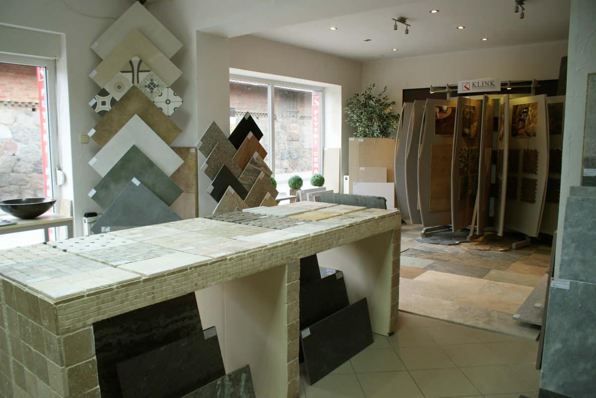Ekspozycja różnorodnych płytek podłogowych i ściennych w salonie sprzedaży, prezentująca wzory, kolory i faktury, z blatem wykończonym mozaiką i ekspozytorem z logo Klink w tle.