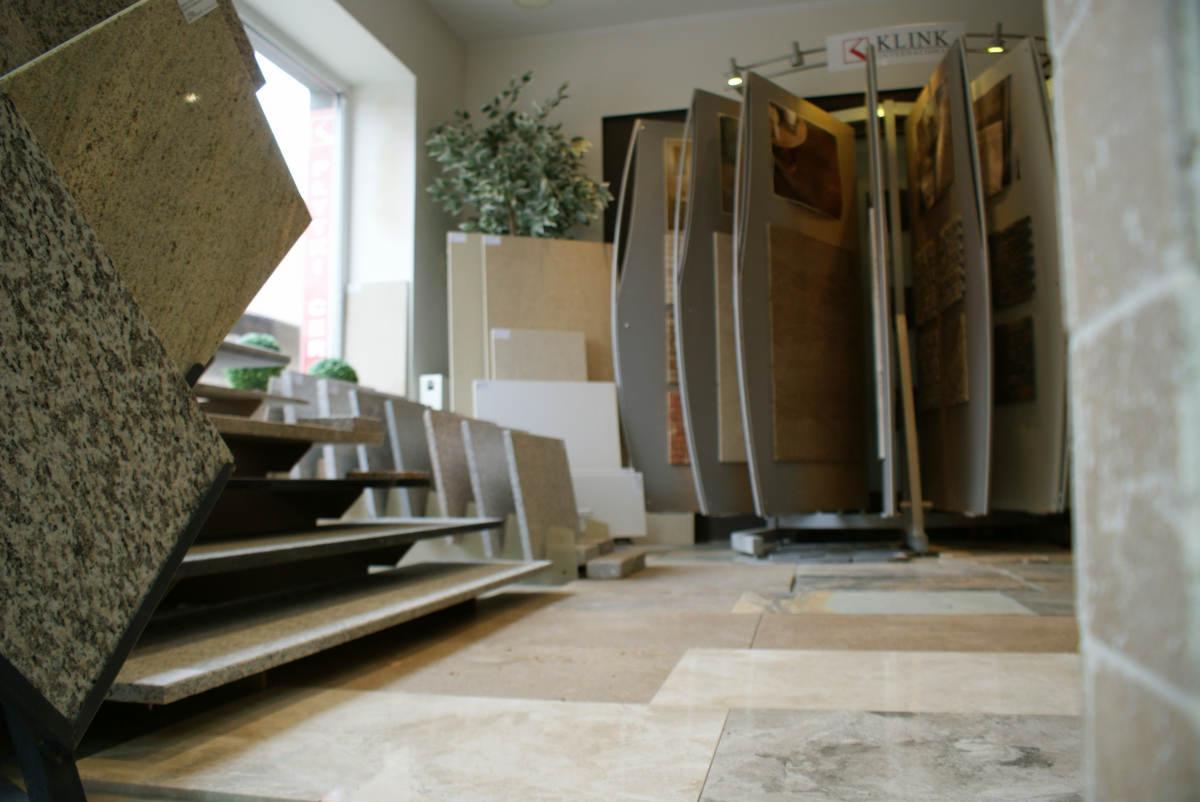 Ekspozycja próbek kostki granitowej w salonie, widoczne różne wzory i kolory, w tle stojak obrotowy z panelami prezentacyjnymi i logo firmy Klink.