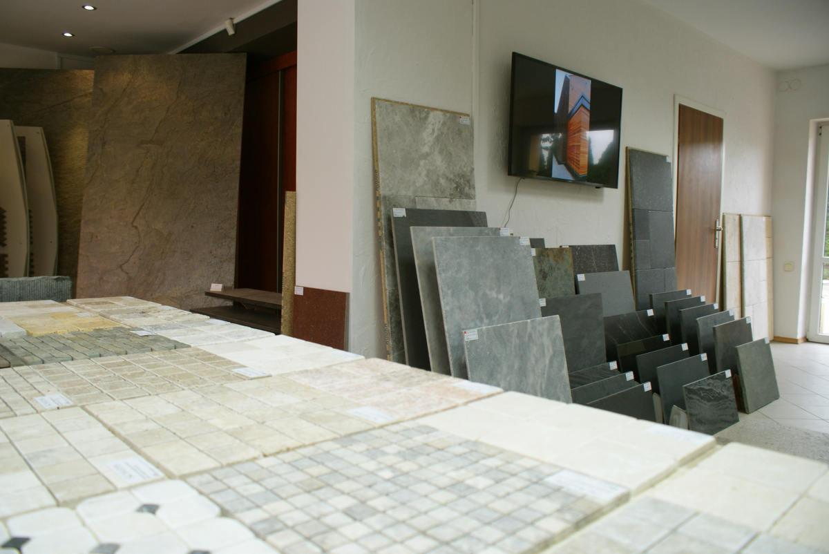 Ekspozycja różnych rodzajów i kolorów kamiennych płytek podłogowych i ściennych w salonie sprzedaży, z widocznymi etykietami cenowymi.