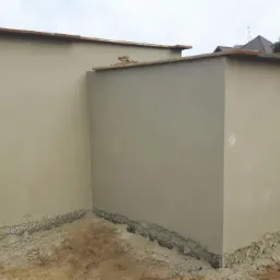 Świeżo otynkowane ściany zewnętrzne budynku gospodarczego z widocznymi śladami zaprawy u podstawy, w tle domy jednorodzinne i pochmurne niebo.