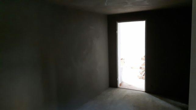 Wnętrze pokoju w trakcie remontu: ściany pokryte świeżym tynkiem maszynowym, surowy beton na podłodze, widok na jasne pomieszczenie przez otwarte drzwi.