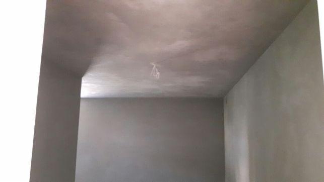 Wnętrze pokoju po nałożeniu szarej warstwy wykończeniowej na ścianach i suficie, widoczne kable wystające z sufitu w miejscu oświetlenia.