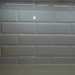 Białe, błyszczące, prostokątne płytki ułożone w rzędach na ścianie, imitujące cegiełki, z delikatnym odbiciem światła.