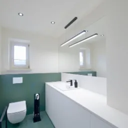 Nowoczesna łazienka z zielonymi heksagonalnymi płytkami na podłodze i ścianie, biała szafka z umywalką i czarną armaturą, minimalistyczne oświetlenie liniowe i lustro.