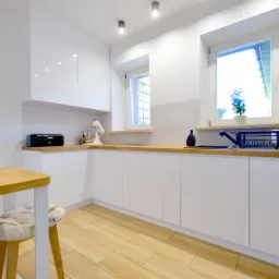 Jasna kuchnia z białymi frontami szafek, drewnianym blatem, drewnianą podłogą i stołem z krzesłami z motywem kwiatowym, oświetlona światłem dziennym i punktowymi lampami sufitowymi.