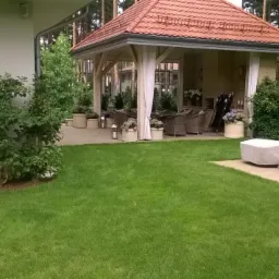 Zadbany trawnik przed domem z altaną ogrodową i zakrytymi leżakami, widoczne drzewa w tle.