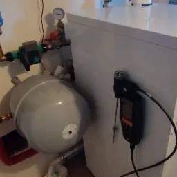 Biały piec grzewczy z podłączonymi rurami, filtrem i manometrem. Widoczny czujnik temperatury przytwierdzony do ściany obok pieca.