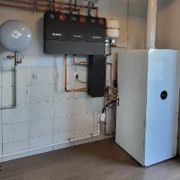 Zainstalowany, nowoczesny system grzewczy Bosch z trzema modułami, naczyniem wzbiorczym i kotłem gazowym kondensacyjnym, z miedzianą instalacją, zamontowany na tle ściany wyłożonej białymi płytkami.