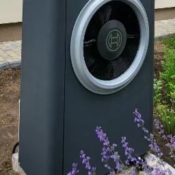 Czarna pompa ciepła Bosch na kamiennym cokole, otoczona kwitnącą lawendą, na tle jasnego budynku z oknami.