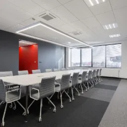 Nowoczesna sala konferencyjna z długim białym stołem, szarymi krzesłami na kółkach, oświetleniem LED w kształcie prostokąta, szarą ścianą z czerwonym akcentem w tle i oknem z żaluzjami.