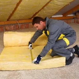 Pracownik w kombinezonie mierzy żółtą wełnę mineralną na poddaszu z ceglaną ścianą i drewnianą konstrukcją dachu.