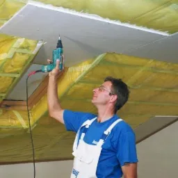 Pracownik w niebieskiej koszulce i białym kombinezonie przykręca płytę gipsowo-kartonową do ocieplonego wełną mineralną poddasza za pomocą wkrętarki.