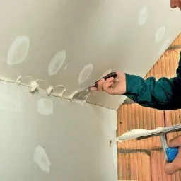 Szpachlowanie sufitu i ściany w narożniku, widoczne nierówności i miejsca po wkrętach, narzędzia w dłoniach wykonawcy, ceglany fragment ściany w tle.