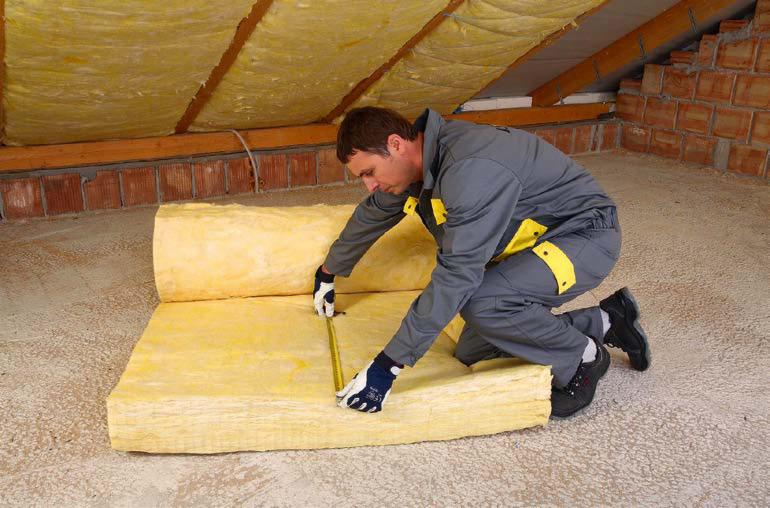 Pracownik w kombinezonie mierzy żółtą wełnę mineralną na poddaszu z ceglaną ścianą i drewnianą konstrukcją dachu.
