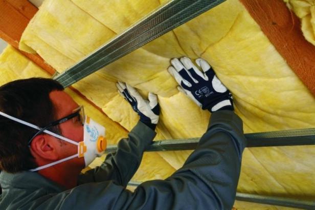 Pracownik w okularach i masce ochronnej, w rękawicach roboczych, montuje żółtą wełnę mineralną pomiędzy profilami metalowymi poddasza.