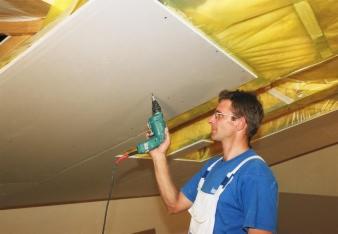 Pracownik w okularach, ubrany w niebieski kombinezon roboczy, przykręca płytę gipsowo-kartonową do drewnianego stelażu na poddaszu za pomocą wiertarko-wkrętarki.