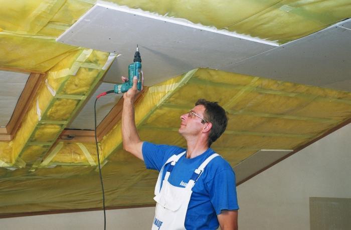 Pracownik w niebieskiej koszulce i białym kombinezonie przykręca płytę gipsowo-kartonową do ocieplonego wełną mineralną poddasza za pomocą wkrętarki.