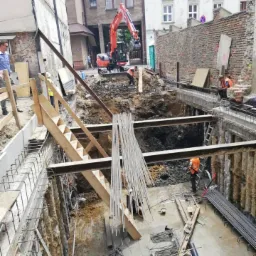 Głęboki wykop pod fundamenty w gęstej zabudowie miejskiej Krakowa, zabezpieczony szalunkiem stalowym, z widoczną koparką i stosem prętów zbrojeniowych. Pracownicy w kamizelkach odblaskowych...