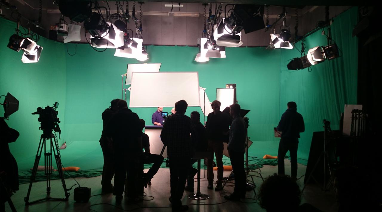 Ekipa filmowa w studio z zielonym tłem, oświetleniem studyjnym i kamerą na statywie, przygotowująca się do nagrania sceny.