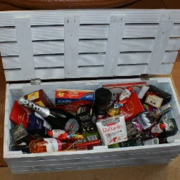 Otwarta, biała, drewniana skrzynia wypełniona różnorodnymi produktami spożywczymi, w tym butelkami alkoholu, czekoladami, kawą i słodyczami, ułożonymi warstwami.