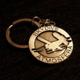 Metalowy brelok z wygrawerowanym motywem skoczka spadochronowego i napisem 'SKYDIVE ATMOSFERA' na ciemnym, teksturowanym tle.