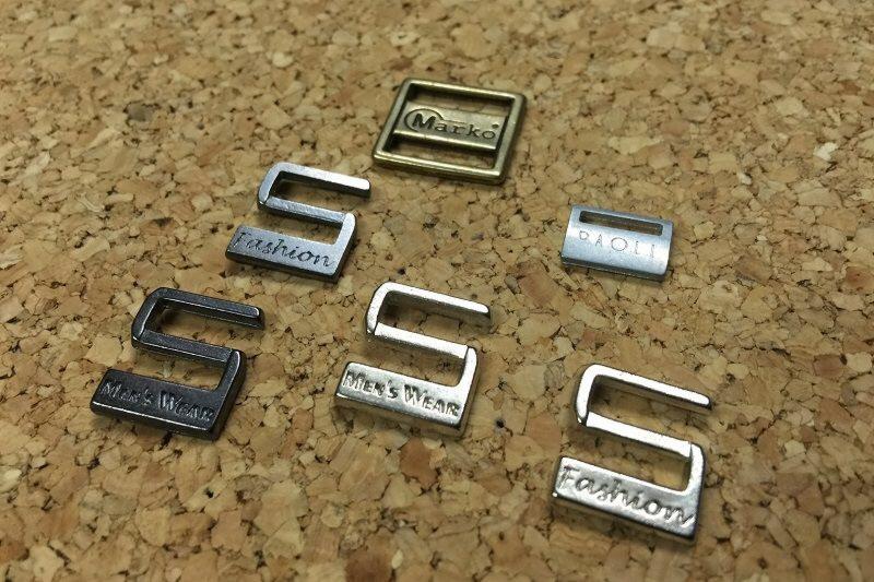 Metalowe klamry i regulatory odzieżowe z napisami 'Marko', 'Fashion', 'Paoli' i 'Men's Wear' na korkowej podkładce.