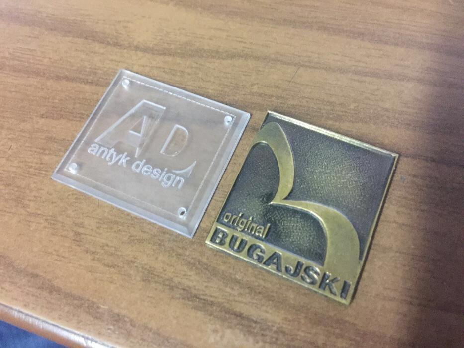 Dwa metalowe emblematy/logo: przezroczysty akryl z napisem 'AD antyk design' i brązowy z napisem 'original BUGAJSKI', leżące na drewnianej powierzchni.