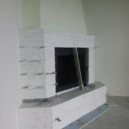 Murowany kominek obłożony białym kamieniem dekoracyjnym w trakcie budowy, widoczne kliny dystansowe i fragmenty płytek podłogowych zabezpieczających posadzkę.