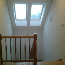 Poddasze z dwoma oknami dachowymi, drewnianą barierką schod&oacute;w i wykończonymi ścianami w jasnym kolorze, widok z g&oacute;ry na schody.