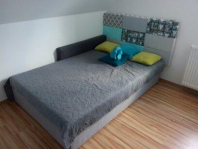 Pokój na poddaszu z szarą sofą z patchworkowym zagłówkiem w odcieniach szarości i turkusu, z żółtymi i turkusowymi poduszkami, na podłodze panele imitujące drewno.