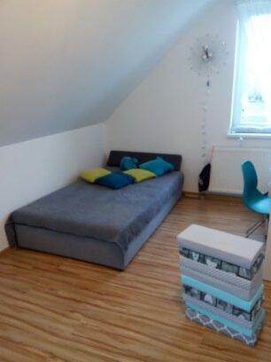 Pokój na poddaszu z łóżkiem, jasnymi panelami podłogowymi i dekoracyjnym zegarem na ścianie, oświetlony przez okno z firanką.