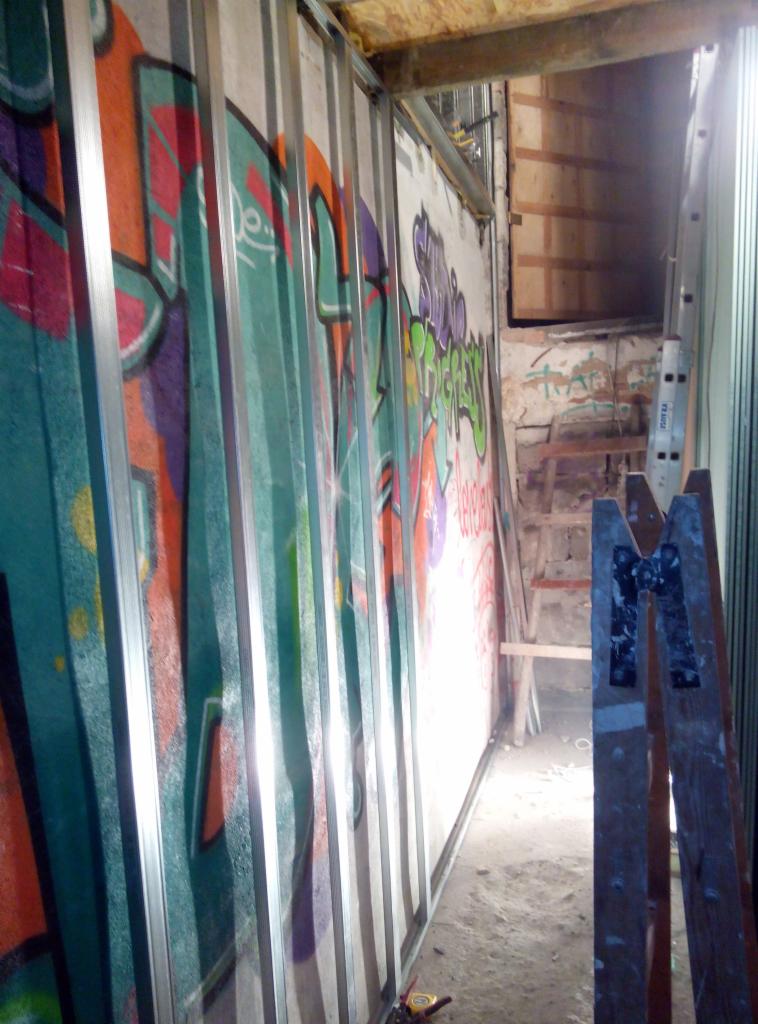 Stelaż z profili metalowych do płyt gipsowo-kartonowych na tle ściany pokrytej graffiti, widoczna drabina i fragment starego muru w głębi pomieszczenia.