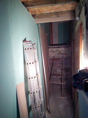 Korytarz w trakcie remontu: ściana wyłożona zieloną płytą gipsowo-kartonową, oparta o nią aluminiowa drabina, w tle stara drewniana drabina i fragmenty odsłoniętej cegły.