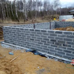 Wznoszenie ścian fundamentowych z szarego bloczka betonowego na placu budowy, widoczne wystające pręty zbrojeniowe, w tle worki z zaprawą i brzozowy las.