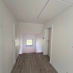 Mariusz Chlipała - Wnętrze poddasza po remoncie: świeżo pomalowane białe ściany, ułożone panele podłogowe imitujące drewno, widoczne drzwi i okno w ścianie szczytowej, oraz klapa rewizyjna w suficie.