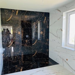 Mariusz Chlipała - Wykończona łazienka z czarno-złotą płytką imitującą marmur na jednej ścianie oraz białą płytką z szarymi żyłami na pozostałych ścianach i podłodze, widoczne okno z białym parapetem.