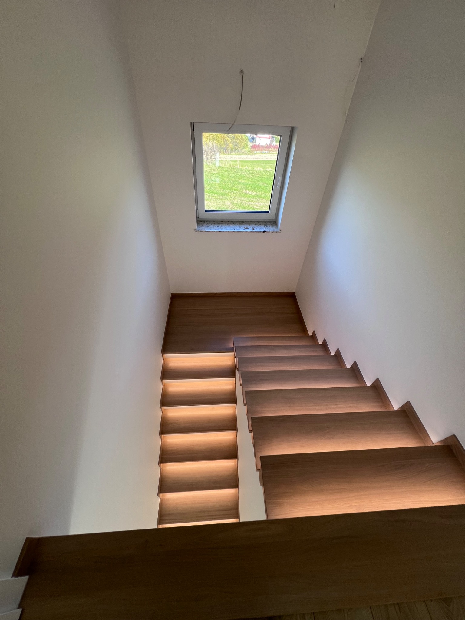 Nowoczesne schody drewniane z podświetleniem LED, widok z góry, małe okno dachowe w tle, minimalistyczne białe ściany.
