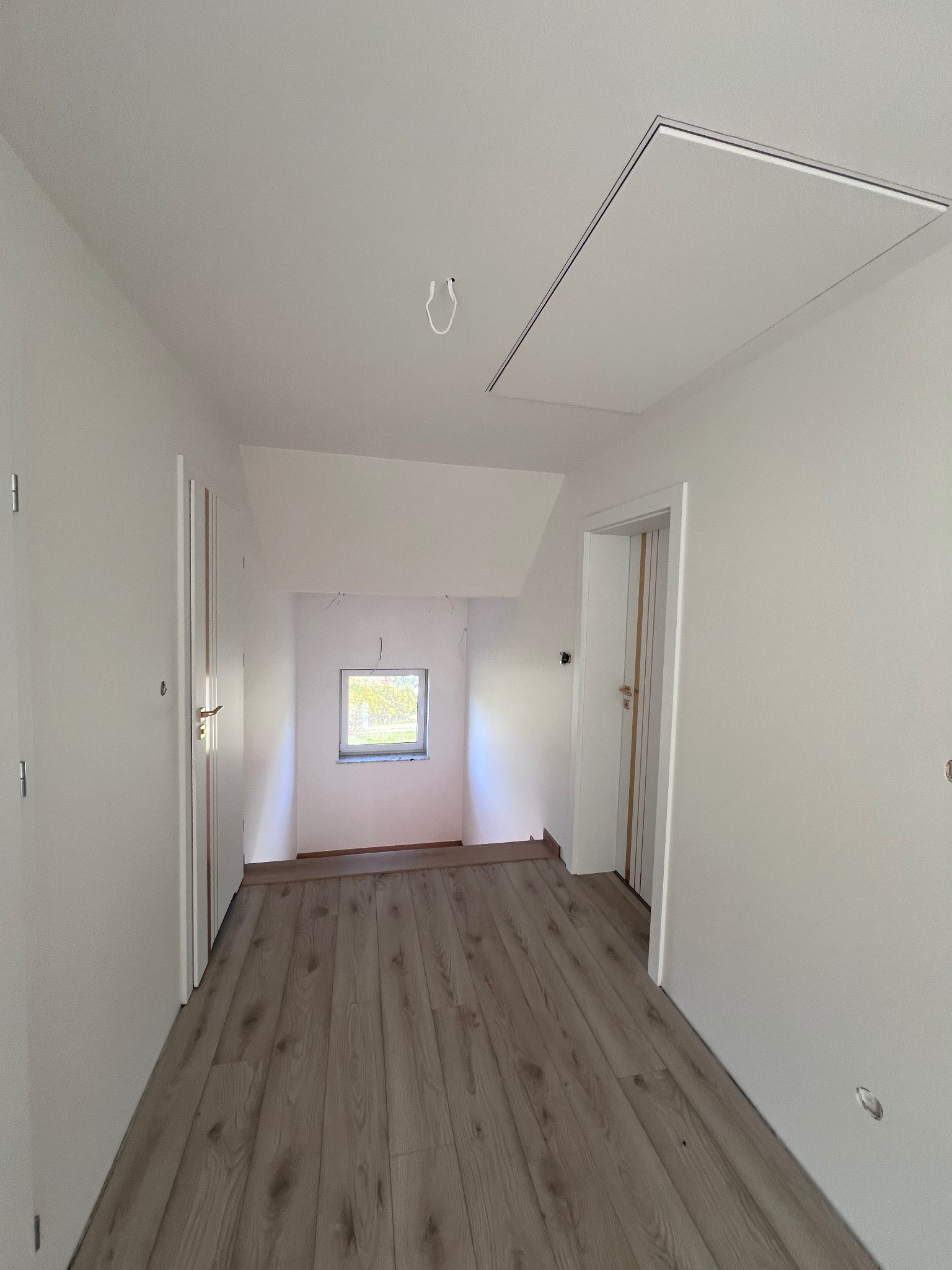 Wnętrze poddasza po remoncie: świeżo pomalowane białe ściany, ułożone panele podłogowe imitujące drewno, widoczne drzwi i okno w ścianie szczytowej, oraz klapa rewizyjna w suficie.