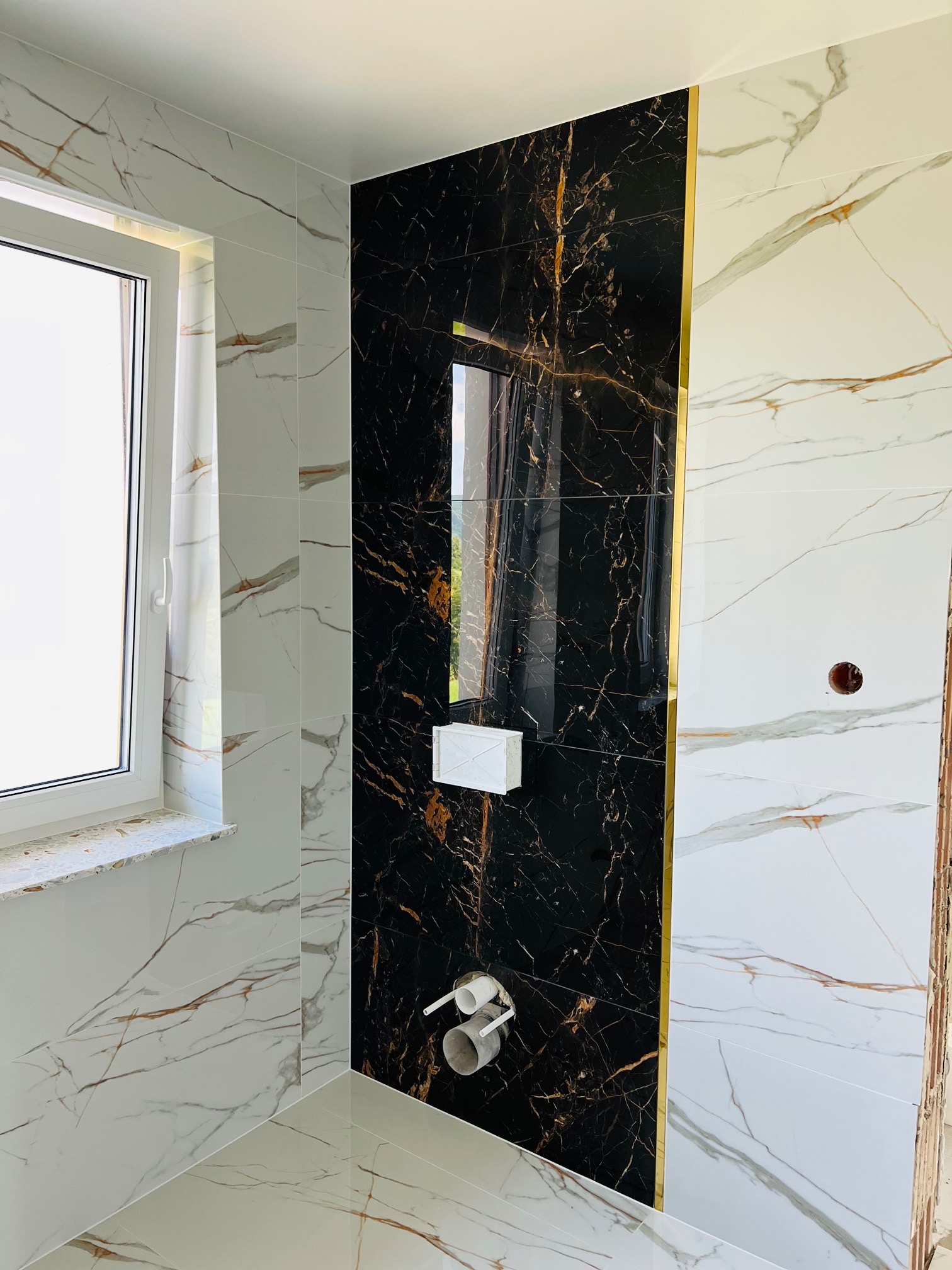 Wykończona łazienka z czarno-białymi płytkami imitującymi marmur, czarna ściana z brokatowym złotym paskiem, biała podtynkowa spłuczka, widoczne rury odpływowe, okno z białymi ramami.