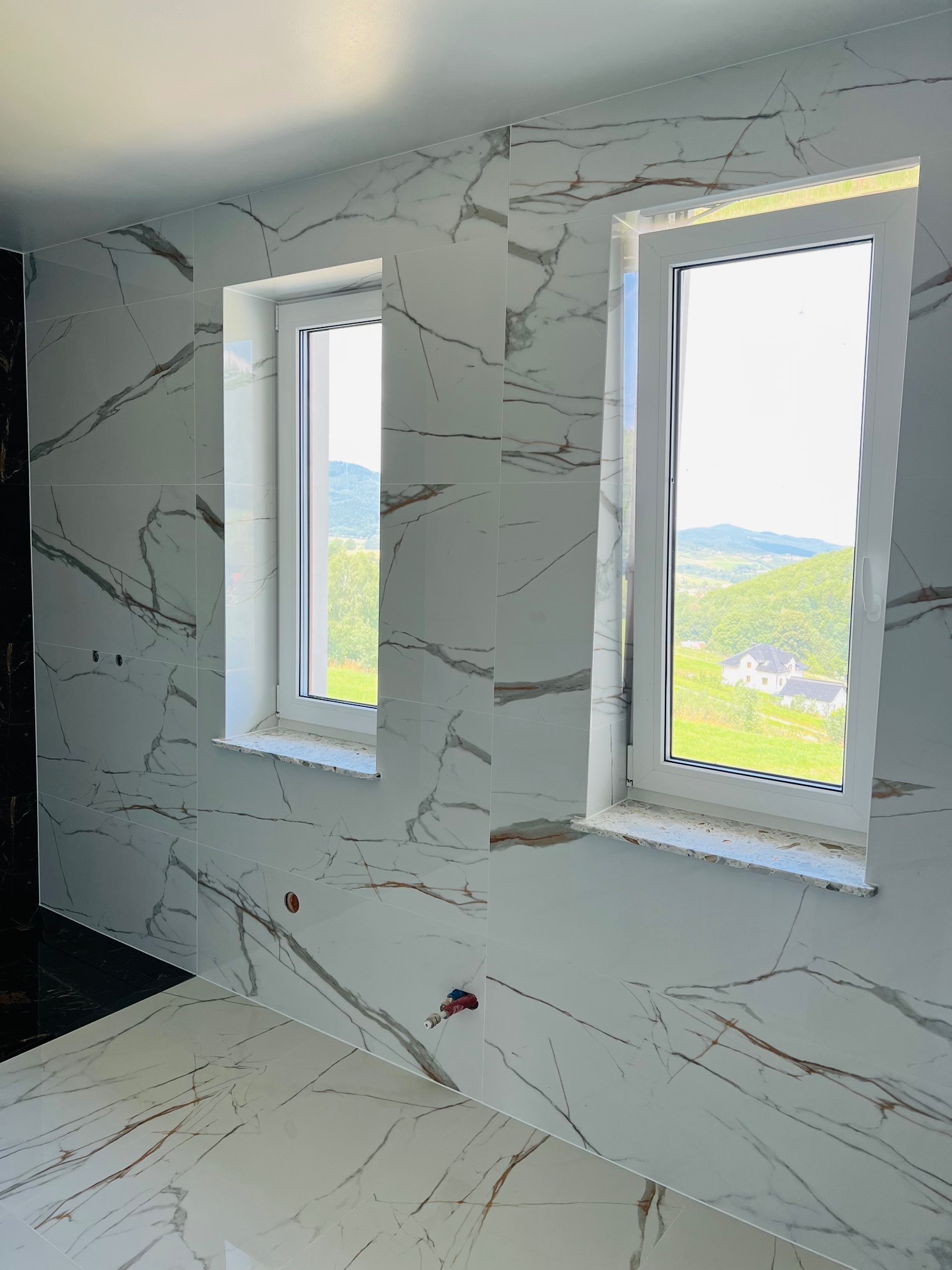 Wykończona łazienka z marmurowymi płytkami, widok na góry przez okna z białymi ramami, instalacja wodna wystaje ze ściany.
