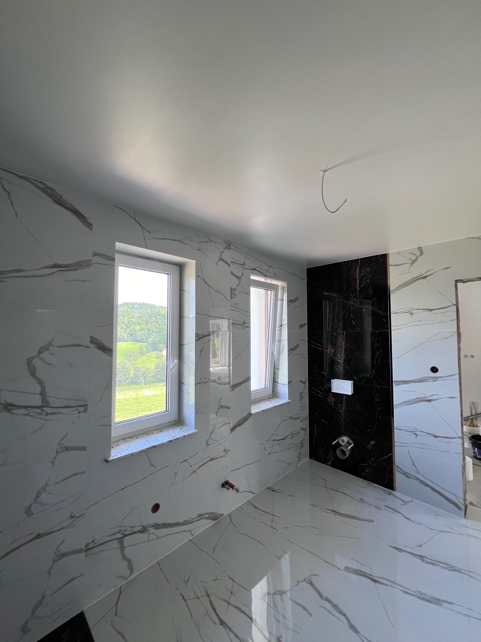 Łazienka w trakcie wykańczania z białymi płytkami imitującymi marmur z brązowymi żyłami, czarna ściana z zabudowanym stelażem WC, widok z okna na zielone wzgórza.