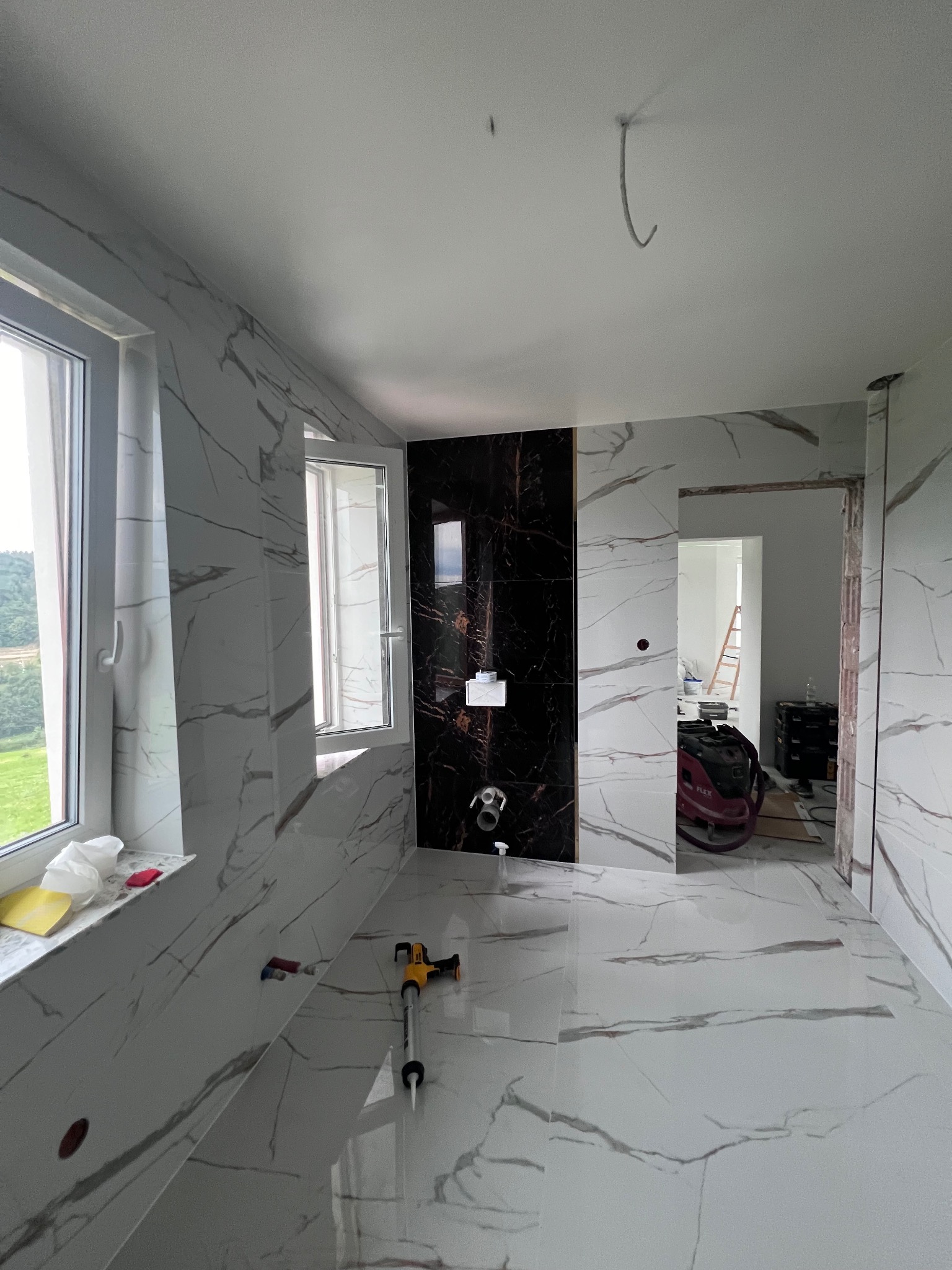 Wnętrze łazienki w trakcie remontu, ściany i podłoga wyłożone białymi płytkami z szarym marmurkowym wzorem, czarna ściana z zamontowaną armaturą, widoczne narzędzia glazurnicze i odkurzacz...