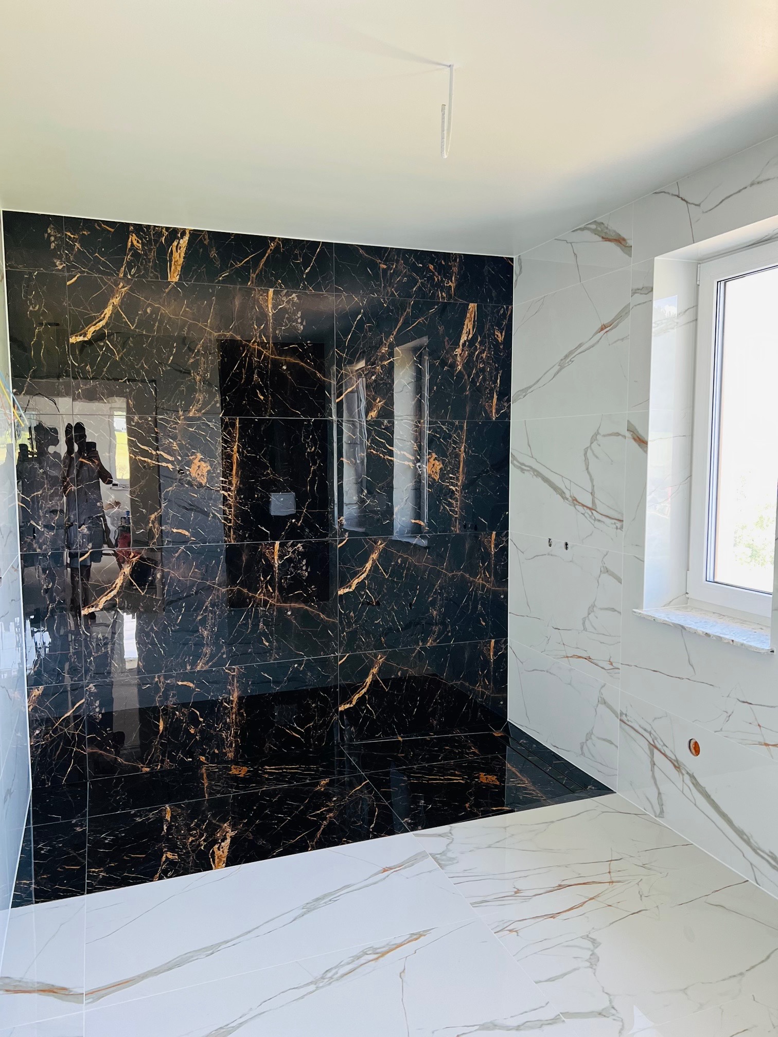 Wykończona łazienka z czarno-złotą płytką imitującą marmur na jednej ścianie oraz białą płytką z szarymi żyłami na pozostałych ścianach i podłodze, widoczne okno z białym parapetem.