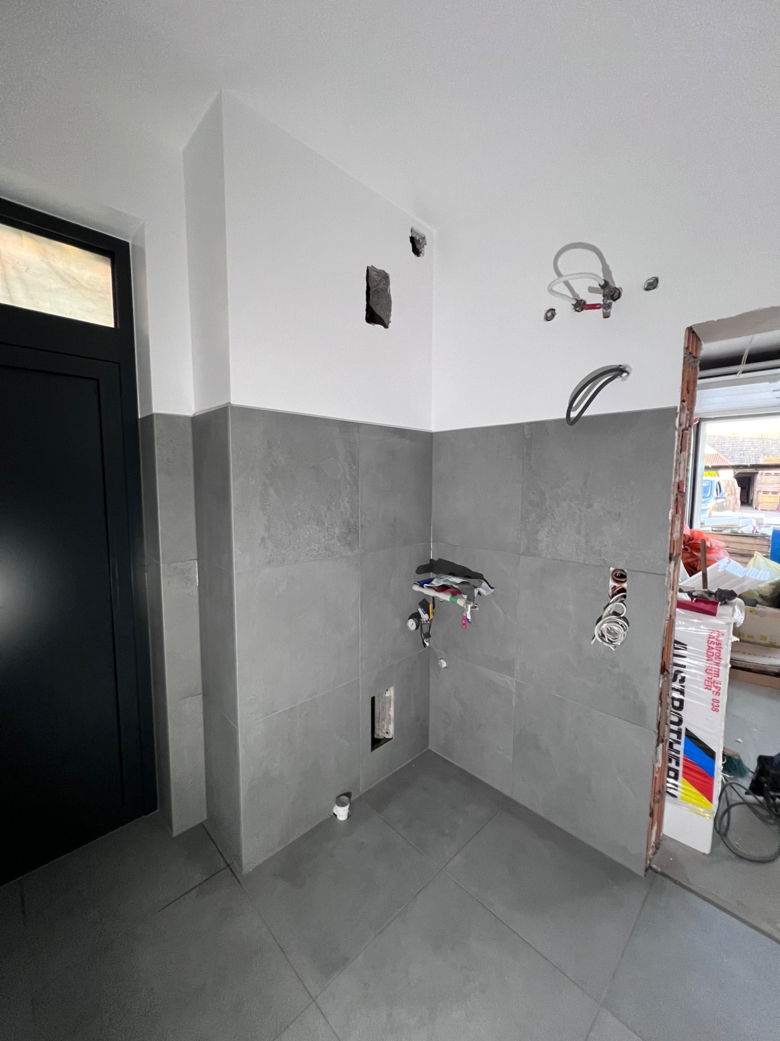 Wnętrze łazienki w trakcie remontu: szare płytki na ścianach i podłodze, widoczne instalacje hydrauliczne i elektryczne, czarne drzwi po lewej, paczka styropianu przy wejściu po prawej.